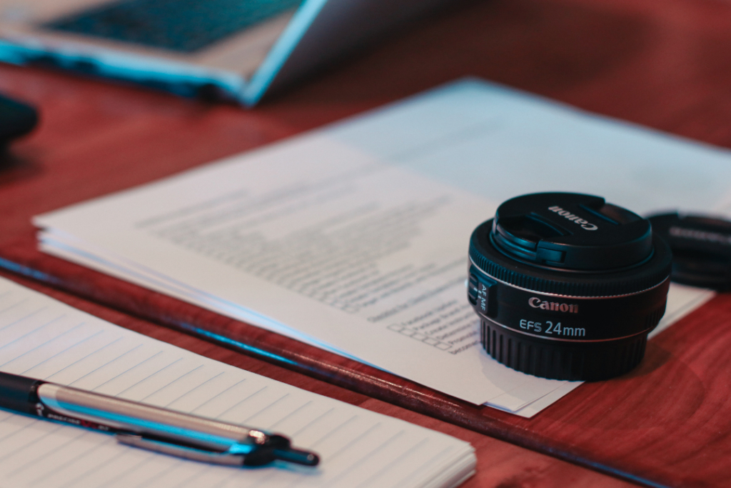 A checklist placed on a brick color desk along with a Canon EFS 24mm lens and a pen and a notebook