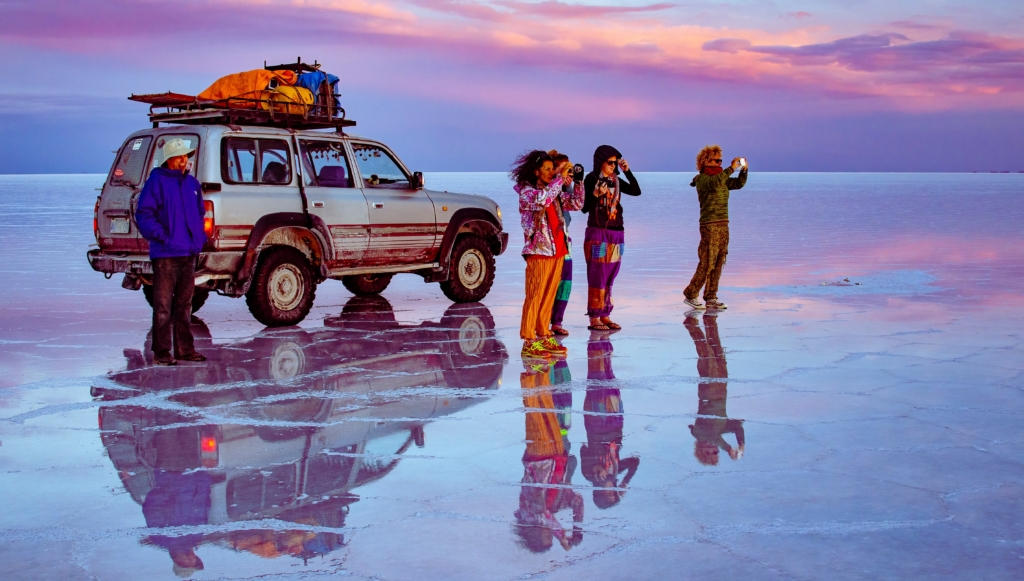 Five people standing in front of a traveler van on a frozen surface using their cameras to capture something