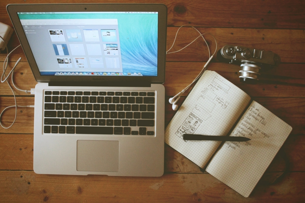 Top shot of an open Macbook along with a camera, notebook and pen | Imagely A laptop, a diary, and a camera placed on top of a desk