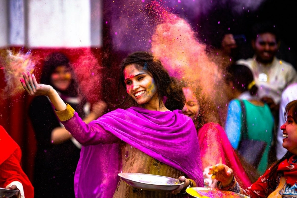 A woman in a colorful sari throwing colored powder into the air and smiling.